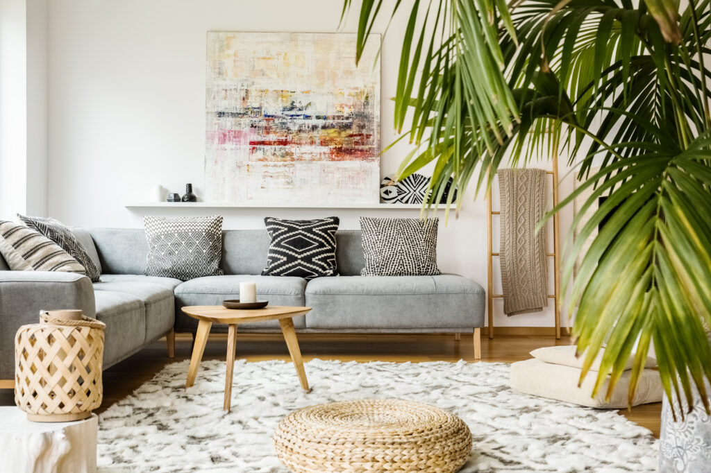 A modern living room with a gray sectional sofa, patterned cushions, a round wooden coffee table, a woven pouf, a large plant, abstract wall art, and a fluffy white rug on a wooden floor.