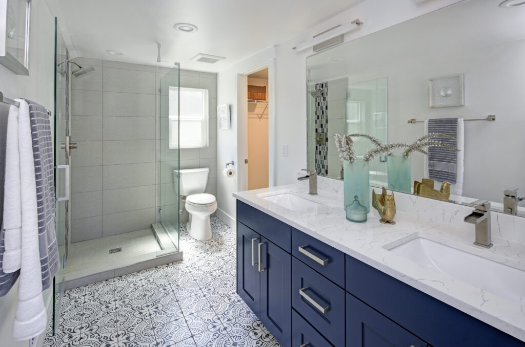 Modern bathroom with patterned tile floor, blue double-sink vanity, white countertop, large mirror, glass shower enclosure, toilet, and decorative vases on the counter. Towels are neatly hung on the wall.