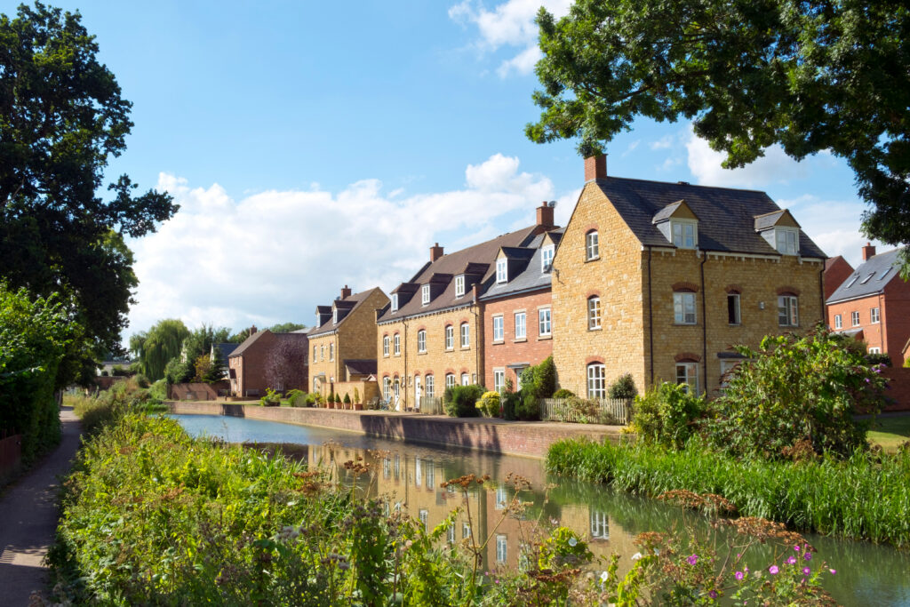 Row of modern brick houses with gabled roofs beside a calm canal, lush greenery and wildflowers in the foreground, and a clear blue sky with scattered clouds above.