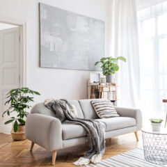 Bright living room with a light gray sofa, striped and solid cushions, a throw blanket, potted plants, a wire coffee table, wooden flooring, and large windows with white curtains; dining area visible in background.