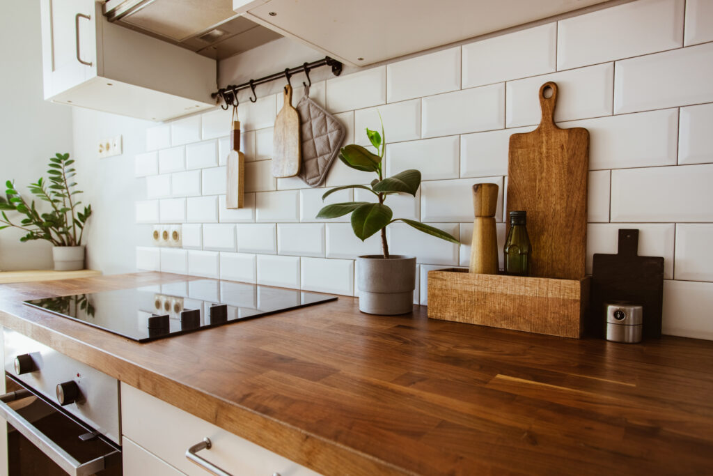 A modern kitchen with a wooden countertop, white subway tile backsplash, potted plants, cutting boards, a stovetop, and kitchen utensils neatly arranged.