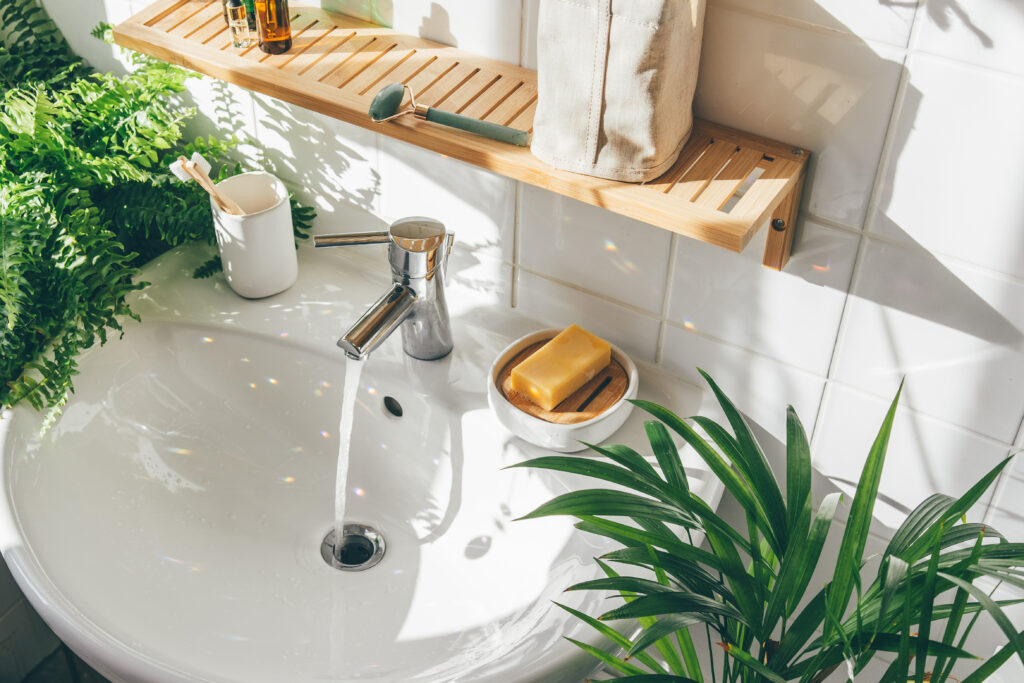 A white bathroom sink with water running, surrounded by green plants, a soap bar in a dish, a cup with toothbrushes, and a wooden shelf holding toiletries against white tiled walls. Sunlight creates sparkles.