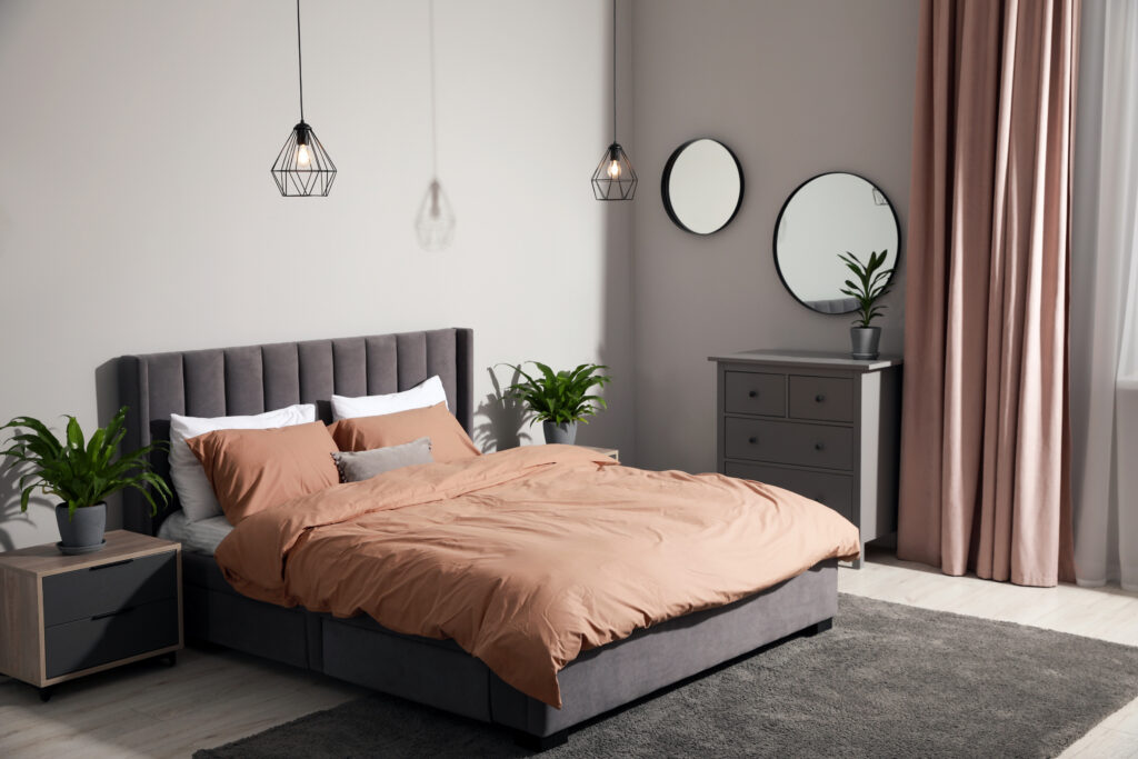 Modern bedroom with a gray upholstered bed, tan bedding, two bedside tables with potted plants, two pendant lights, a dresser with mirrors, and pink curtains by a window. The room is softly lit and neatly organized.
