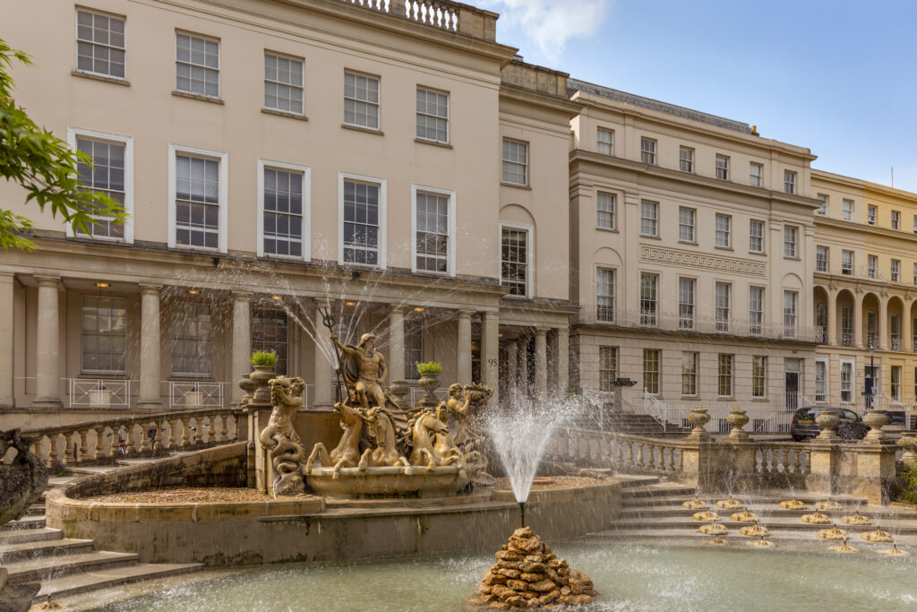 A grand stone fountain with statues and water jets in front of a large, elegant neoclassical building with tall windows, columns, and balconies under a blue sky.